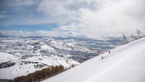 Skiers carve fresh powder on a wide-open slope at Deer Valley Resort, surrounded by snow-covered mountains and winter skies in Utah’s Wasatch range