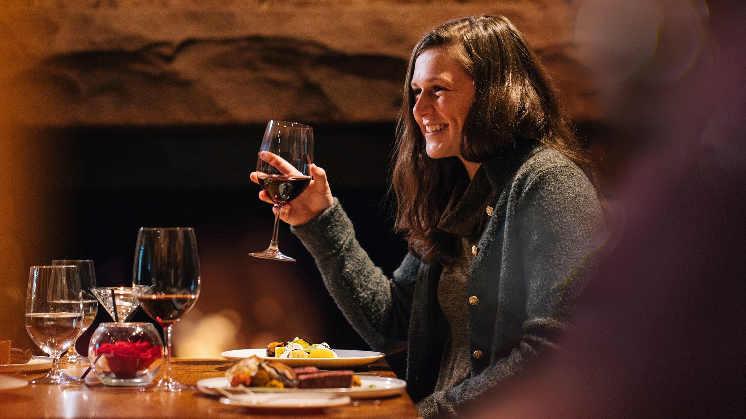 Guest drinking wine and smiling while sitting next to fireplace.