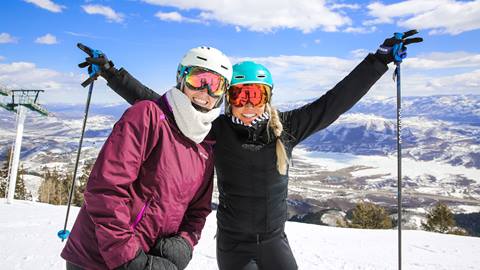 Two women wearing ski gear are posing for a picture with mountains in the background.