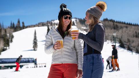 Two women standing outside drinking beer while wearing ski gear.