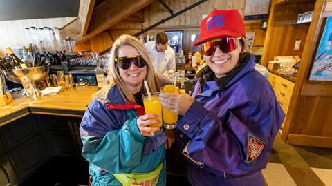Two guests wearing retro ski gear holding drinks at the Sticky Wicket bar.